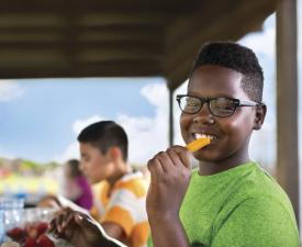 his selected focused image is of a smiling boy eating a piece of cheese, with two other children eating in the background.